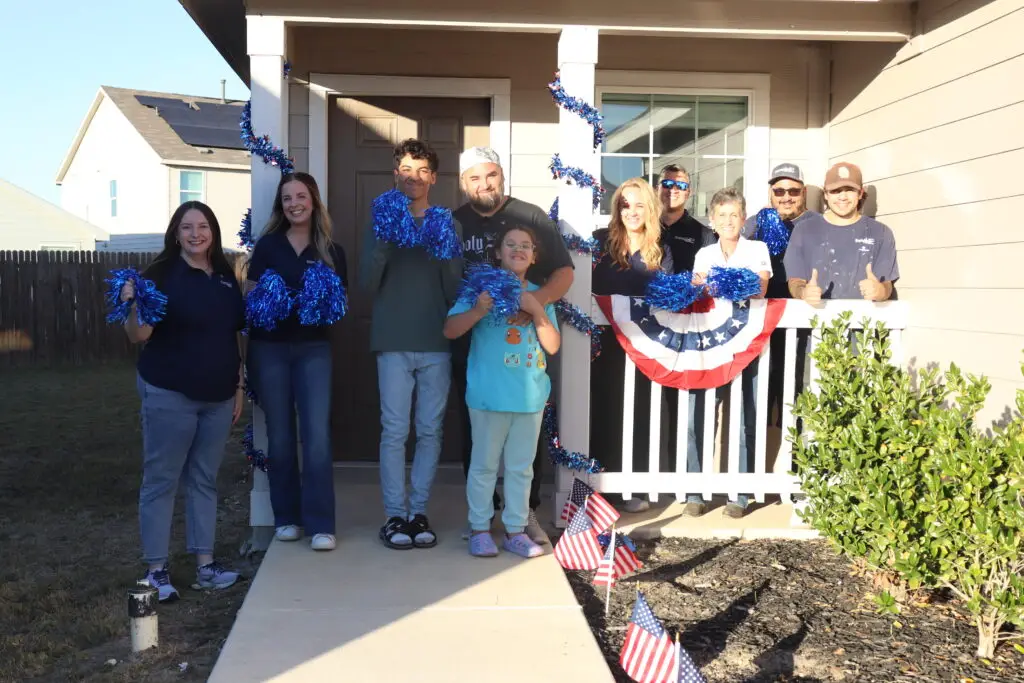 Group of eleven people on a porch, smiling and holding blue pom-poms, with red, white, and blue decorations and small American flags nearby.